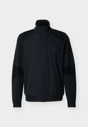 Veste noire à fermeture éclair intégrale avec col montant, poignets et ourlet côtelés, poches zippées, et logo discret "Porsche BOSS" sur la poitrine.