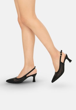 Gambe con tacchi slingback intrecciati neri con punta appuntita e tacchi curvi di media altezza su uno sfondo chiaro uniforme.