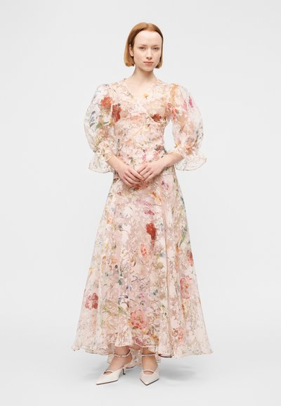 Robe longue en tissu transparent à motif floral avec manches bouffantes, encolure en V et base rose pâle ; associée à des chaussures de couleur claire.