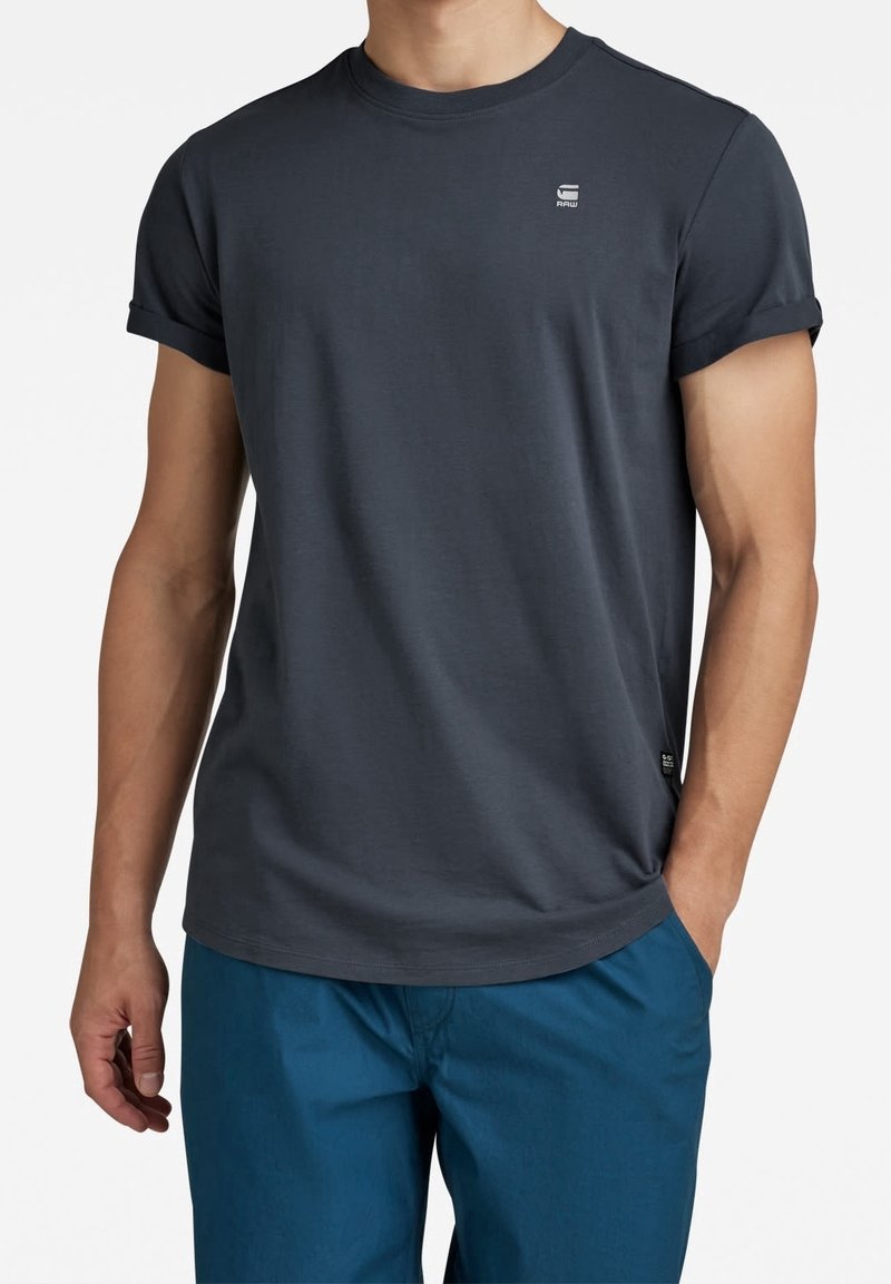 Homme portant un t-shirt gris foncé à manches courtes avec un petit logo sur la poitrine et un pantalon bleu, debout avec une main dans la poche.