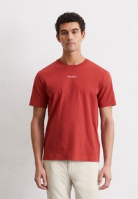 Red cotton t-shirt featuring a crew neck and short sleeves. Logo printed in white at the chest. Worn with light beige pants.