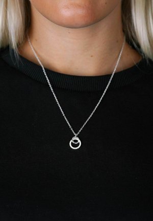 Silverhalsband med en cirkulär berlock som har ett texturerat band och en slät inre ring, hängande från en fin kedja på en svart topp.
