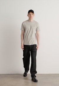 Camiseta gris de manga corta con logo, combinada con pantalones cargo sueltos negros y zapatos de cuero negros. El modelo lleva una gorra negra.