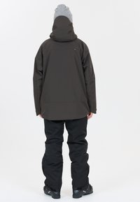 Waterdichte donkerbruine jas met een capuchon, gecombineerd met zwarte broek. De jas heeft een ontspannen pasvorm en een gladde textuur, perfect voor buitenshuis.