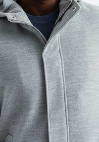 Chaqueta gris con cremallera y capucha, con una textura suave, cuello entallado y una capa interior en contraste de color azul marino.