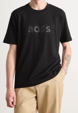 Junge Person trägt ein schwarzes T-Shirt mit dem "BOSS"-Logo in linierter Schrift, beige Hosen und ein Schlangen-Tattoo auf dem rechten Unterarm.
