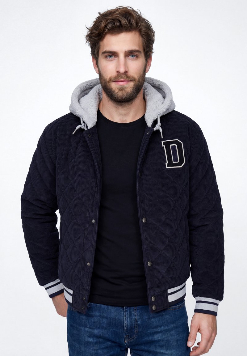 Homme aux cheveux bruns et barbe, portant une veste matelassée bleu marine avec une capuche grise et un patch en forme de lettre "D", une chemise noire et un jean bleu, debout devant un fond blanc.