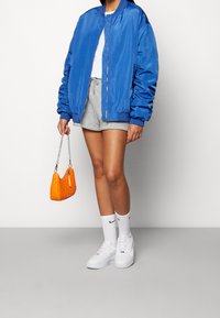 Chaqueta bomber azul, pantalones cortos grises, camiseta blanca y zapatillas blancas. Sosteniendo un bolso acolchado naranja con una cadena.