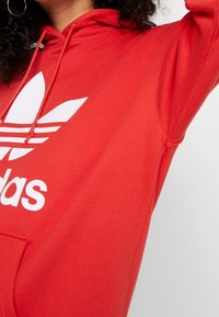 Kvinna i en röd Adidas-hoodie med vitt logo, lyfter en arm, visar lockigt hår och gyllene hoopörhänge.