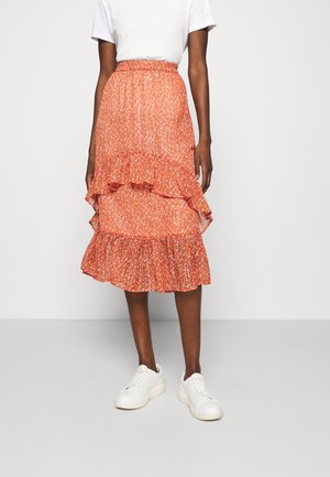Une personne porte une jupe midi fleurie orange taille haute avec des volants, un haut blanc à manches courtes et des baskets blanches, debout sur un sol gris.