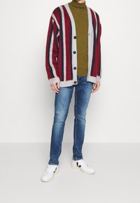 Punainen ja harmaa raidallinen neuletakki vihreän poolopaidan päällä, yhdistettynä sinisiin farkkuihin ja valkoisiin lenkkareihin, joissa on mustia yksityiskohtia. Villa materiaalia, napitettu.