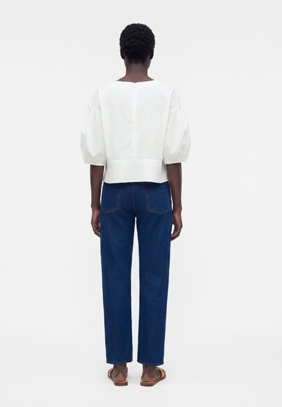 Άτομο με κοντά σγουρά μαλλιά που φοράει λευκό τοπ με φουσκωτά μανίκια, σκουρομπλε τζιν με ίσια γραμμή και καφέ επίπεδα σανδάλια, στέκεται με την πλάτη.