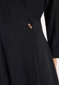 Czarna sukienka z teksturowanej tkaniny, z kopertowym krojem i pojedynczym brązowym guzikiem po boku. Długie rękawy o luźnym kroju.