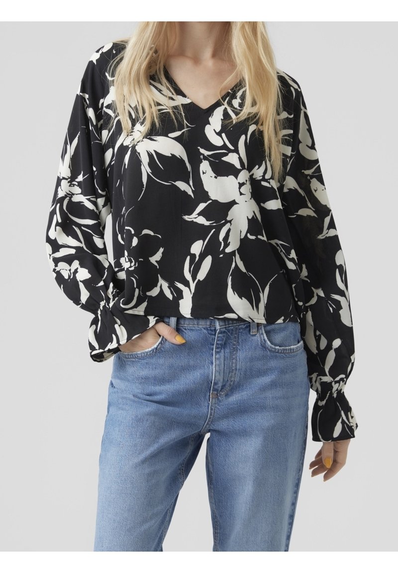 Femme portant un chemisier noir à manches longues avec un imprimé floral blanc et un jean bleu clair, une main dans la poche, sur fond uni.
