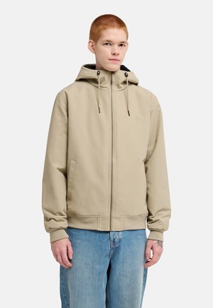 Jeune personne aux cheveux roux courts portant une veste à capuche beige zippée et un jean bleu clair sur un fond uni.