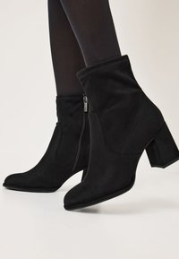 Bottines noires en daim, dotées d'une fermeture éclair sur le côté, d'un bout pointu et d'un talon épais avec une texture lisse.