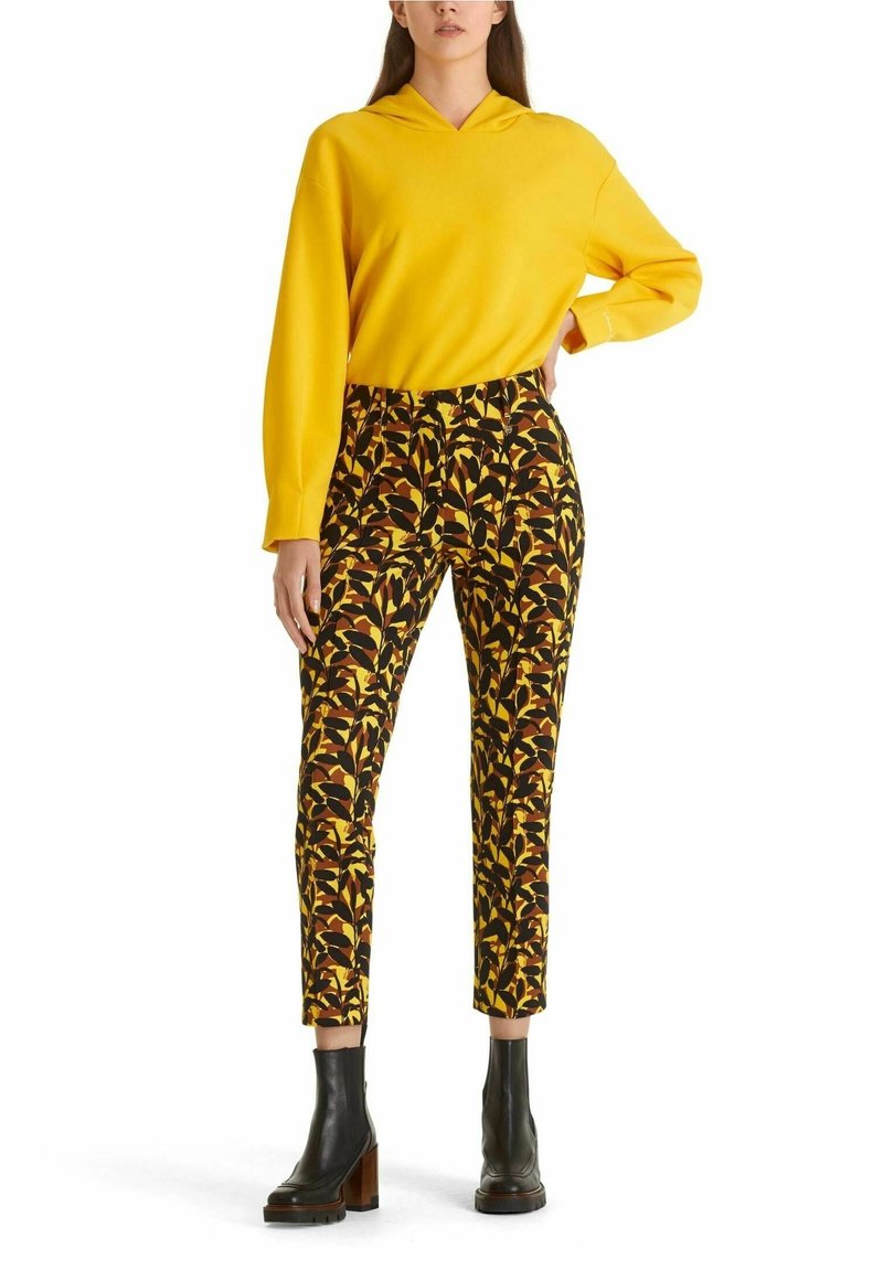 Femme portant un sweat à capuche jaune vif, un pantalon taille haute à motifs avec un design feuillu noir et marron, et des bottines noires à talons.