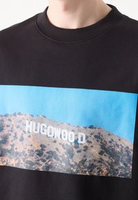Černá mikina s velkým grafickým potiskem kopce a nápisem "HUGOWOOD D" na pozadí modré oblohy. Měkká, texturovaná látka.