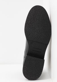 Semelle d'une chaussure habillée noire avec une semelle en caoutchouc texturée et la taille 37 marquée près de la voûte plantaire, sur un fond blanc.