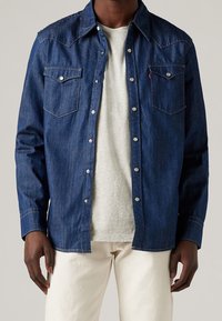 Mörkblå denimskjorta med tryckknappar, spetsig ok-design och två bröstfickor. Bärs över en ljusgrå t-shirt, matchad med ljusa byxor.