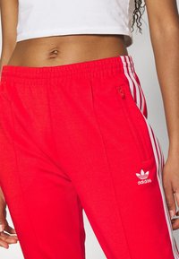 Röda Adidas träningsbyxor med vita sidosträck och en dragkedjeficka. Tillverkade av ett mjukt material med resår i midjan.