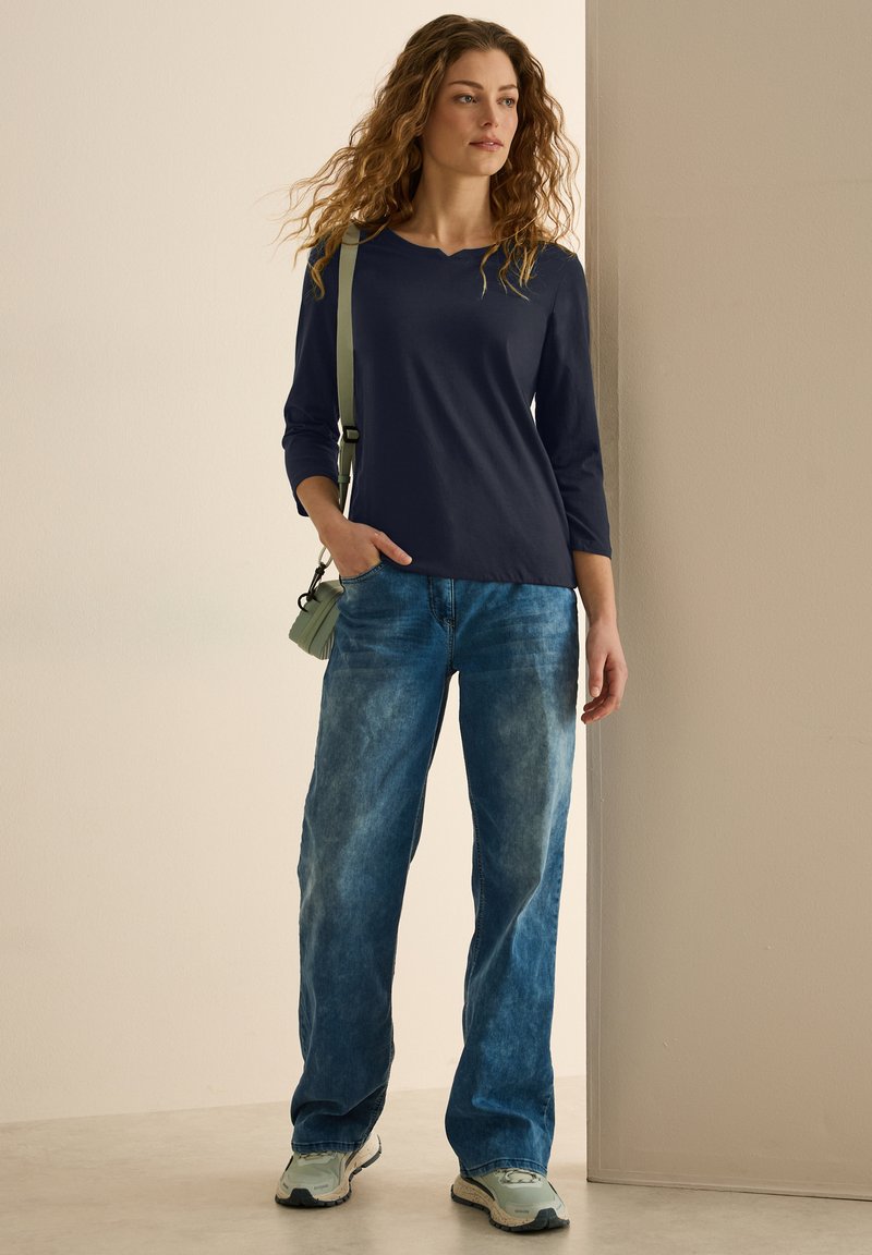 Marinblus med lång ärm, ljusblåa urtvättade jeans och ljusblå sneakers. Skjortan har en rundad halsringning; jeansen är löst sittande med en avslappnad stil.