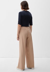 Nainen, jolla on lyhyet ruskeat hiukset, pukeutuneena mustaan kolmannelta neljännekseltä hihatoppiin, beiget leveät housut ja valkoiset tennarit, seisoo valkoista seinää vasten.