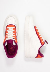 Le sneakers Nike Air Force 1 presentano una tomaia in pelle bianca con dettagli arancioni e viola, una punta perforata e una suola in gomma bianca.