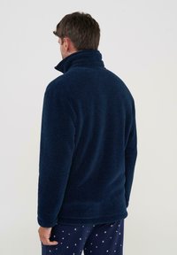 Sudadera de forro polar azul oscuro con un cuello alto y tejido texturizado, mostrada desde la parte posterior, combinada con pantalones de pijama estampados.