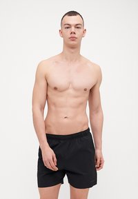 Juodi maudymosi šortai su elastinga juostele ir lygia tekstūra. Modelis stovi priešais paprastą baltą foną, demonstruodamas šortus.