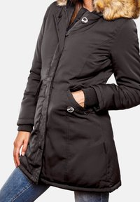 Parka negra acolchada con capucha de piel sintética, acentos de botones plateados, bolsillos laterales y puños acanalados. La tela parece suave y resistente al agua.