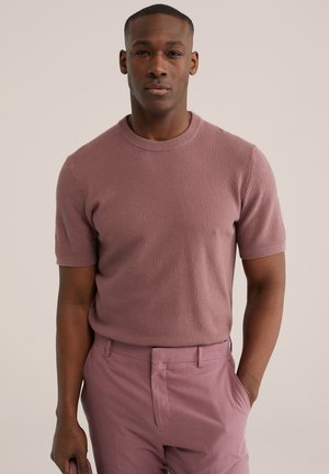 Bărbat purtând un pulover cu mâneci scurte texturat, de culoare mov pal, și pantaloni asortați mov pal, stând cu o mână în buzunar, pe un fundal simplu.