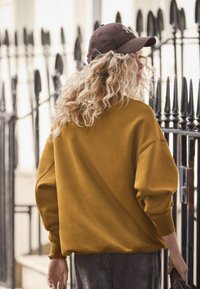 Felpa oversize giallo senape con spalle scese, polsini a costine e tessuto strutturato. Abbinata a un cappello marrone e pantaloni grigi.
