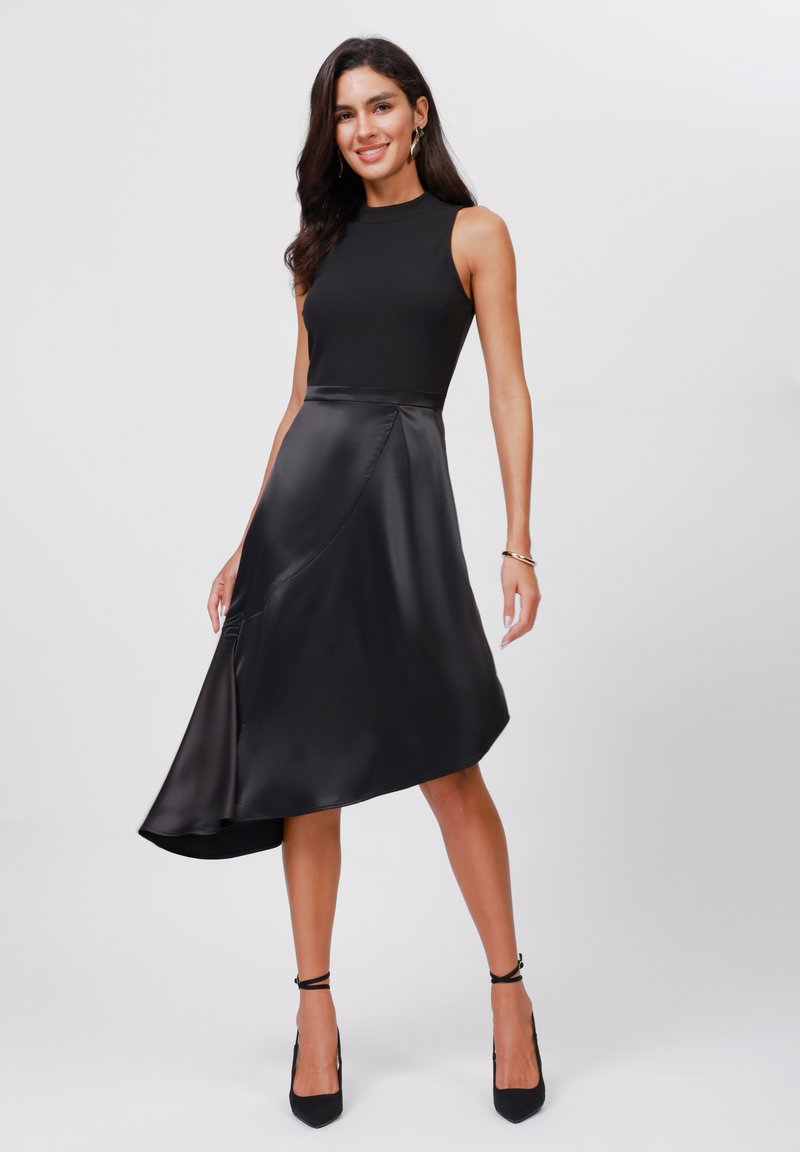 Zwart mouwloos jurk met een aansluitende top en een vloeiende asymmetrische satijnen rok, met een gladde textuur en subtiele glans. Inclusief zwarte hakken.