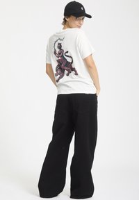 Hvid t-shirt med et farverigt grafisk motiv af en person, der rider på en tiger på ryggen, samlet med løstsiddende sorte jeans. Sort kasket båret bagud.
