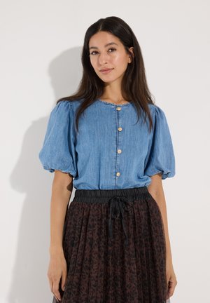 Frau trägt eine blaue Bluse mit kurzen Puffärmeln, goldenen Knöpfen und einen hoch taillierten schwarzen und braunen gemusterten Rock mit Kordelzug.