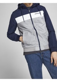 Hooded jacket in navy blue, gray, and white. Features a front zipper, side pockets, ribbed cuffs, and a logo on the chest.