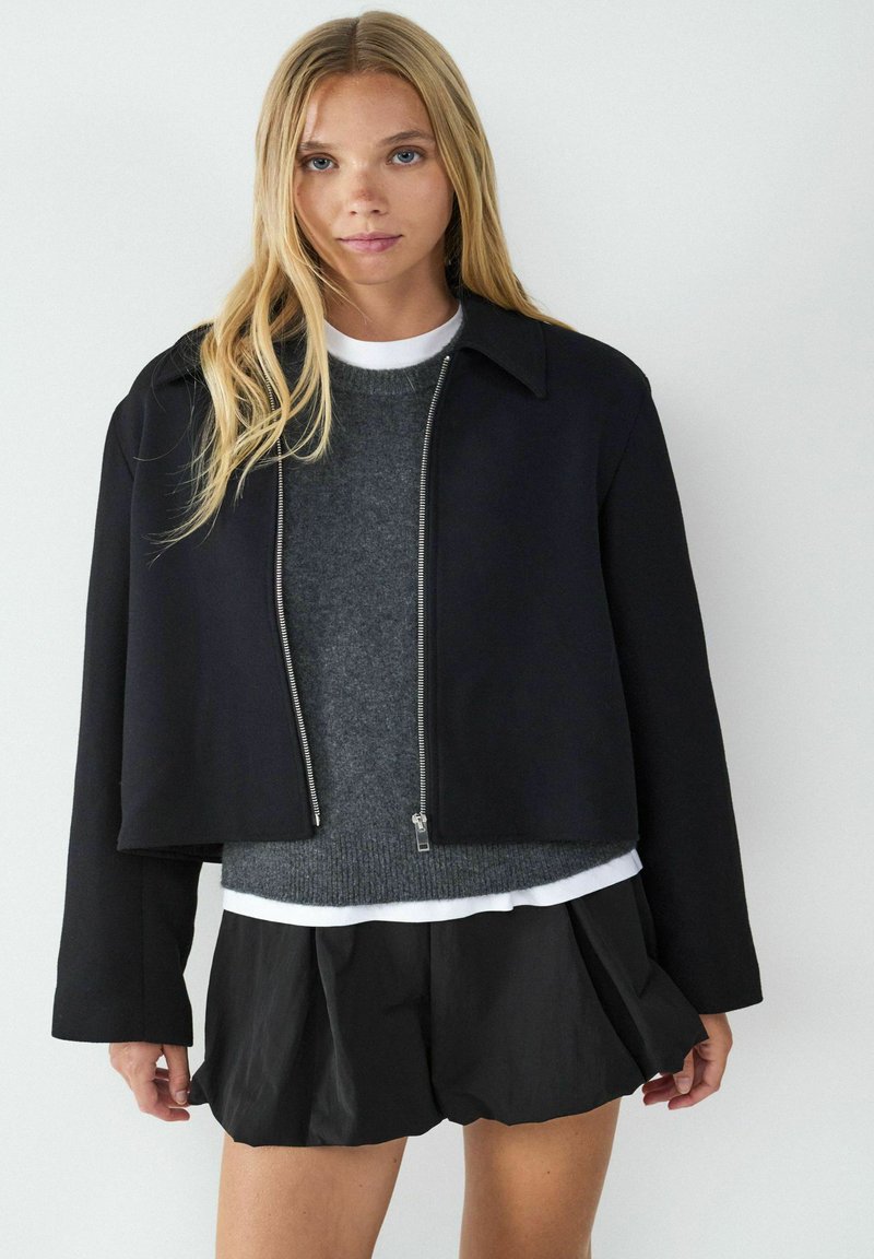 Schwarze, taillierte Jacke mit Reißverschluss und Kragen, kombiniert mit einem grauen Pullover und schwarzen, plissierten Shorts, die subtile Texturen und Schichten aufweisen.