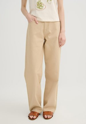 Persona que lleva pantalones beige de pierna ancha, top crema con gráfico y sandalias marrones abiertas en los dedos, de pie con una mano en el bolsillo.
