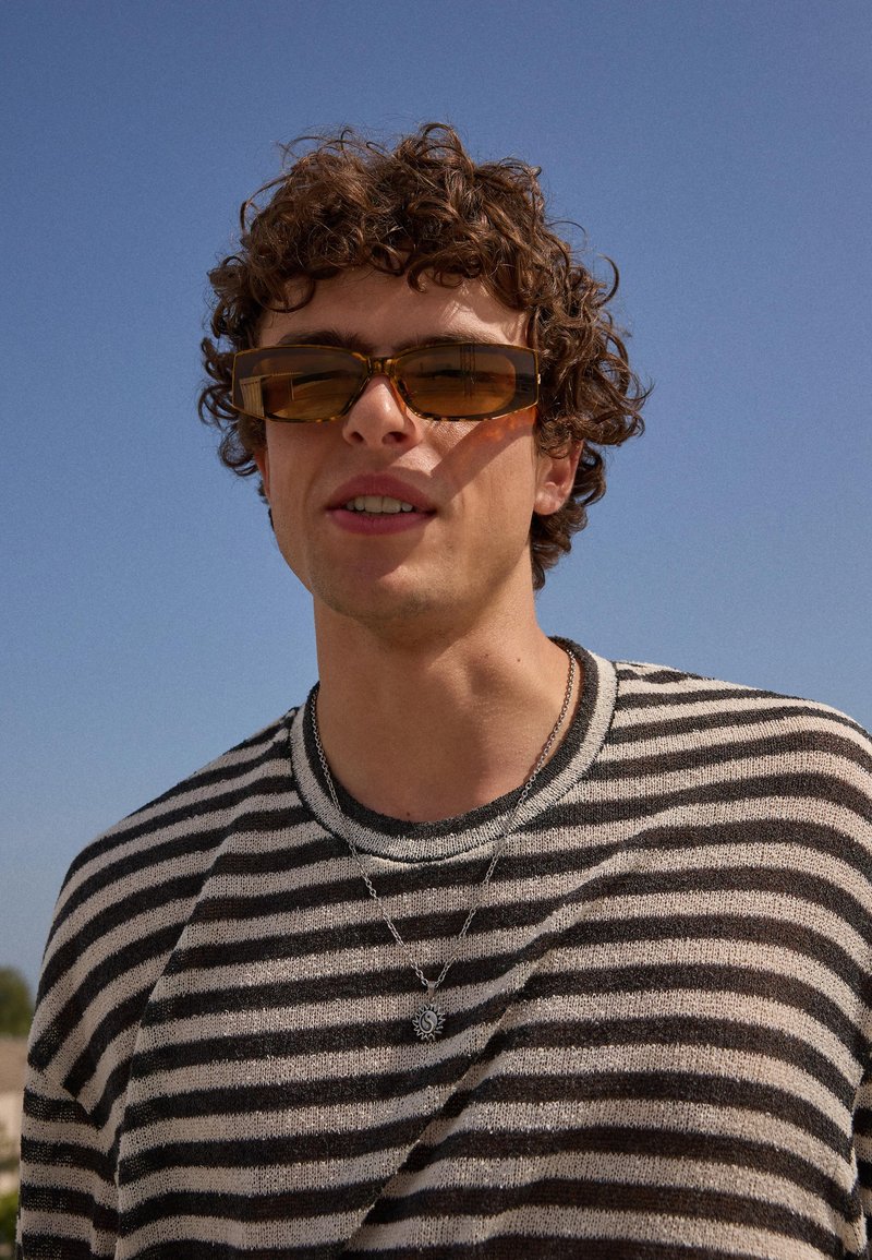 Jeune homme aux cheveux bouclés portant des lunettes de soleil rectangulaires marron, un pull rayé noir et blanc et un collier avec pendentif soleil sous un ciel dégagé.