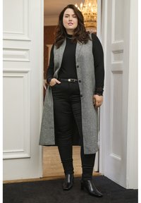 Chaleco largo gris con botones negros sobre un top negro ajustado, combinado con pantalones negros ajustados y botas negras de tobillo, con detalles de encaje.