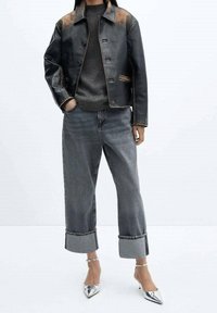 Persoon die een zwart leren jack, een antraciete trui, grijze jeans met brede omslagen en zilverkleurige pumps met puntige neus draagt, staand tegen een witte achtergrond.