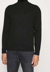 Svart, stickad polotröja med långa ärmar, ribbade muddar och nederkant. Har en subtil logotyp på bröstet, tillverkad av mjukt tyg.