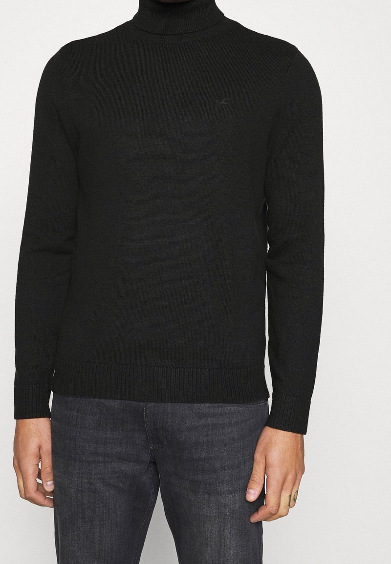 Schwarzer gestrickter Rollkragenpullover mit langen Ärmeln, gerippten Bündchen und Saum. Verfügt über ein dezentes Logo auf der Brust, hergestellt aus weichem Stoff.