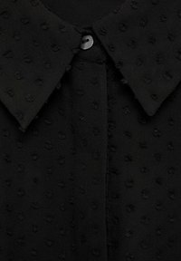 Blouse noire à col en tissu texturé, présentant de petits motifs en relief et une fermeture centrale à boutons.