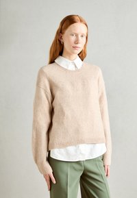 Világos bézs gyapjús jumper laza szabással, bordázott mandzsettákkal és szegéllyel, fehér galléros ing fölött viselve. Az outfitet olíva zöld nadrág egészíti ki.
