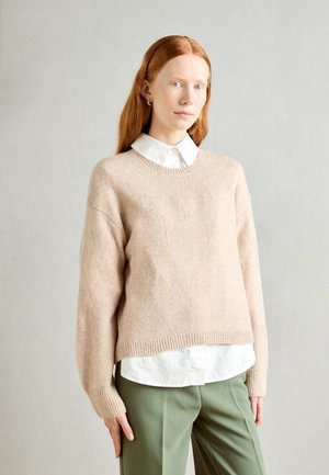 Light beige wool sweater with a relaxed fit, ribbed cuffs, and hem, layered over a white collared shirt. Olive green pants complete the look.