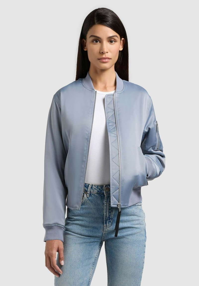 Femme aux longs cheveux bruns portant une veste bomber bleu clair, un haut blanc et un jean bleu, se tenant devant un fond uni.