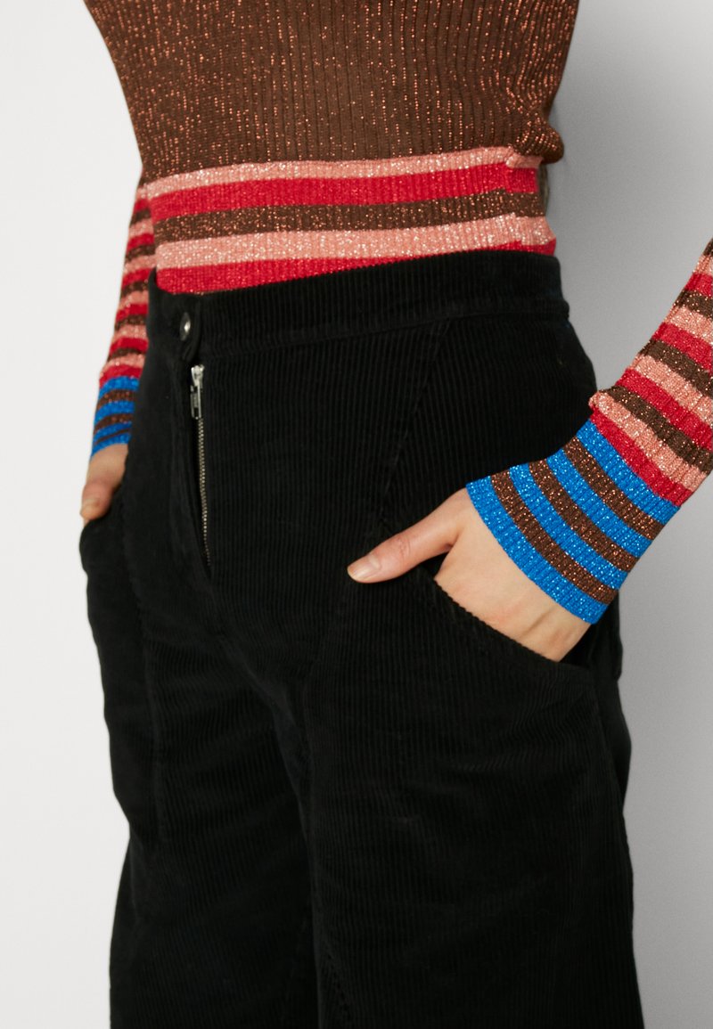Haut à manches longues côtelé, marron, avec des rayures horizontales colorées, associé à un pantalon en velours côtelé noir avec une poche zippée et un tissu texturé.