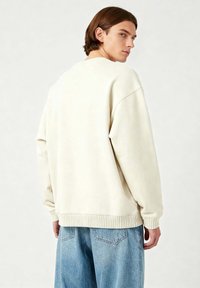 Felpa oversize beige chiaro con polsini e orlo a costine, realizzata in materiale morbido. Abbinata a jeans in denim blu chiaro.